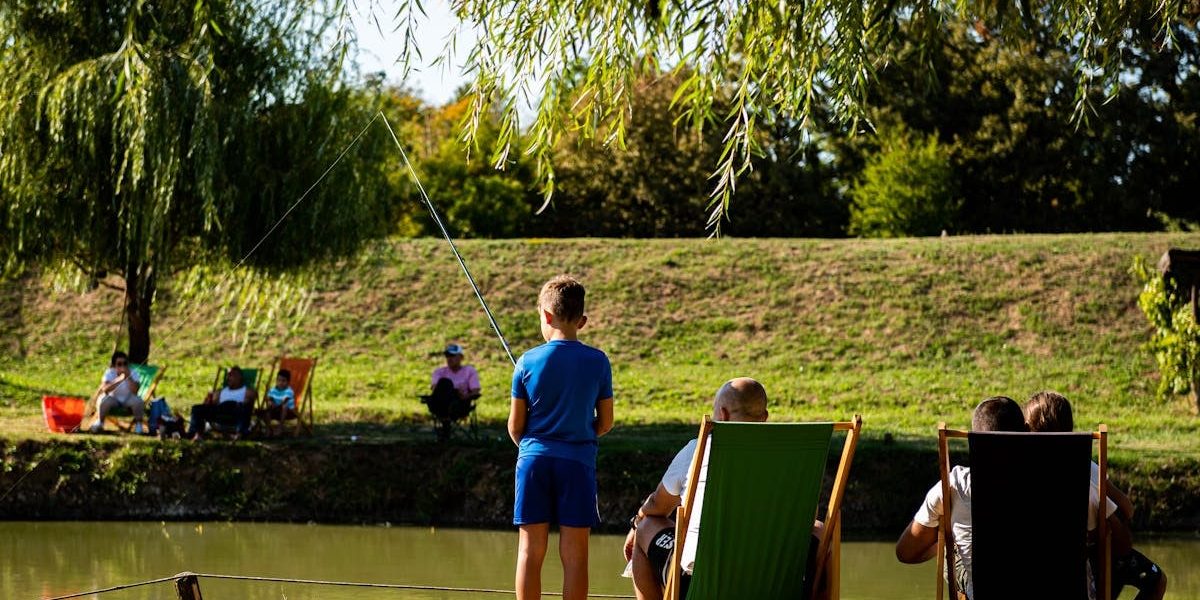 Young boy fishing by a pond on a sunny day, surrounded by trees and relaxed people.