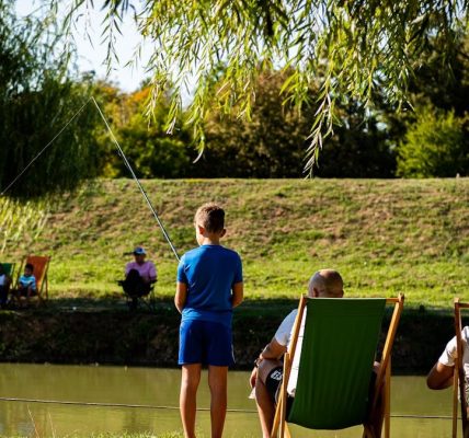Young boy fishing by a pond on a sunny day, surrounded by trees and relaxed people.