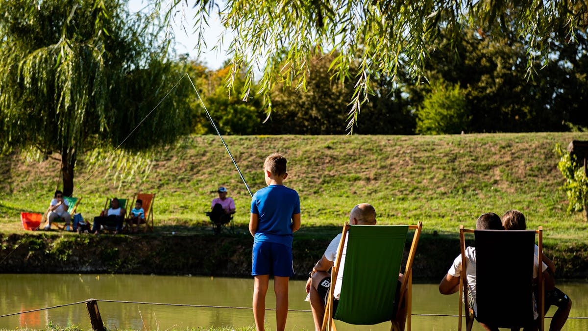 Young boy fishing by a pond on a sunny day, surrounded by trees and relaxed people.